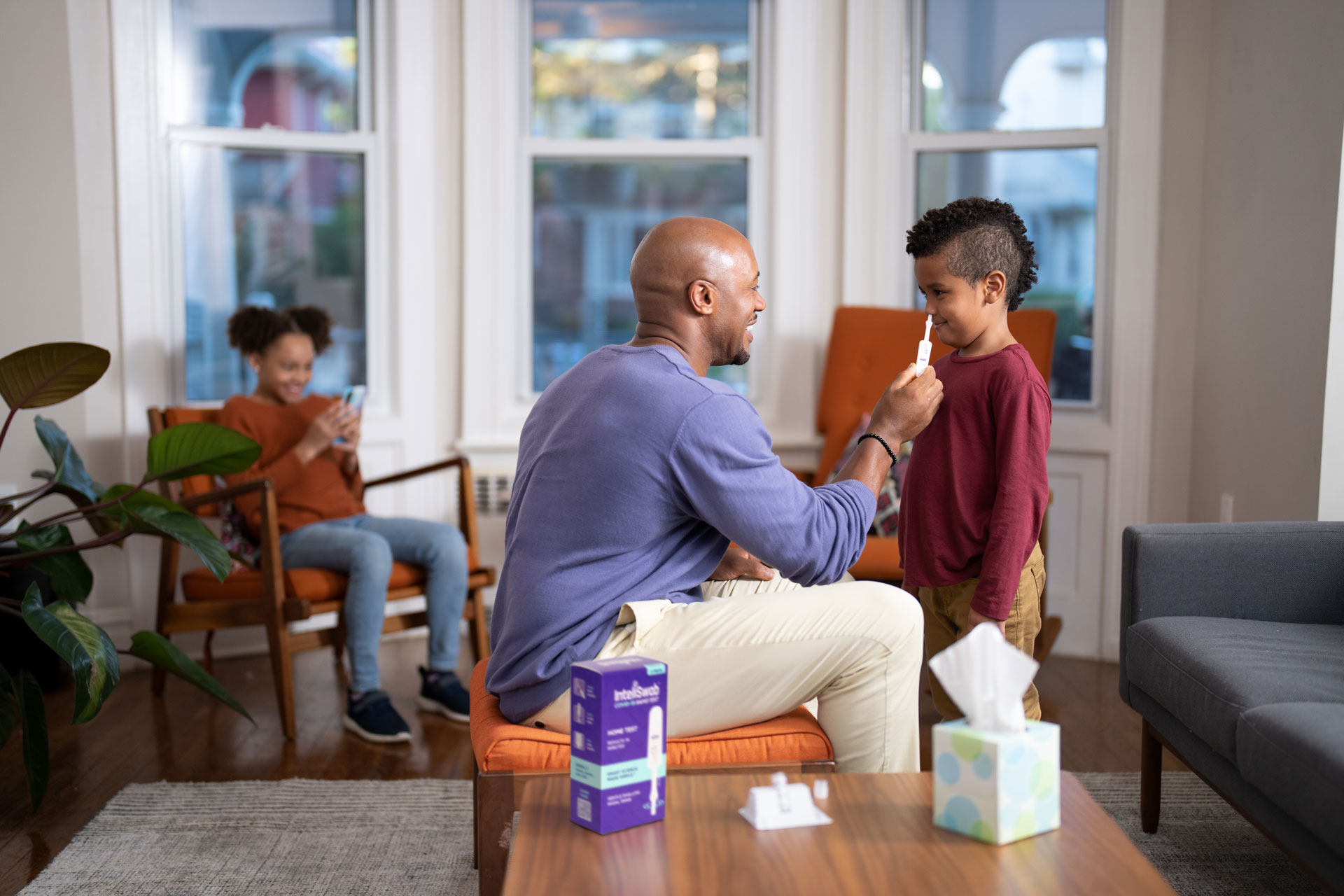 A man swabs a child's nose at home. They are both comfortable and smiling. Another child is smiling while on their phone in the background.