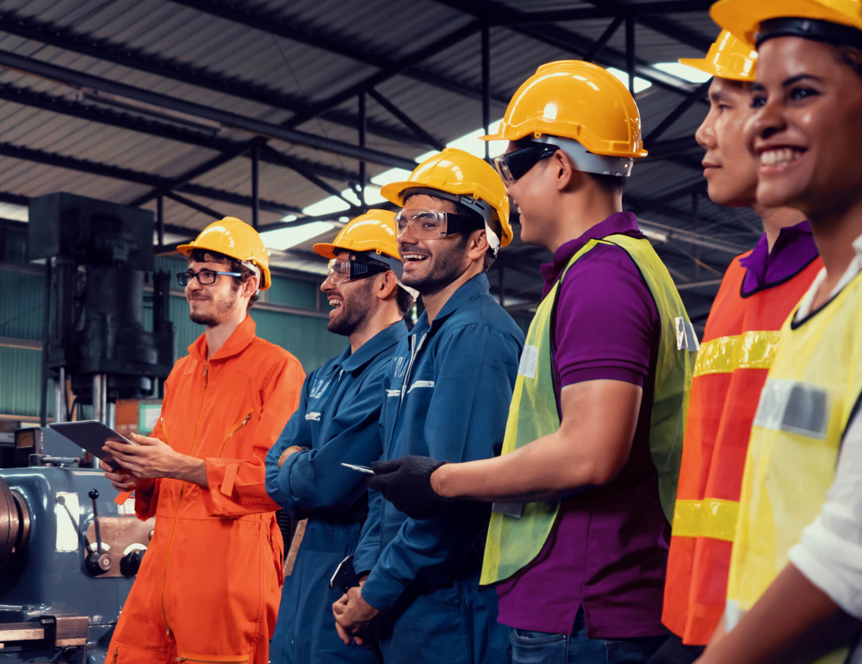 A group of workers stand in a factory wearing hard-hats and safety gear while laughing.